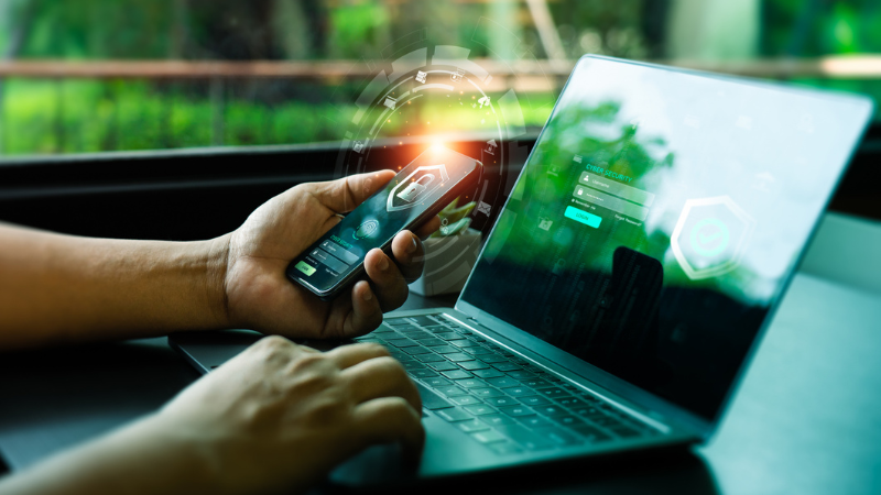 A person using a smartphone and laptop with glowing security icons on both screens, symbolizing secure access and data protection. The image highlights the importance of robust Cybersecurity measures to safeguard sensitive information in a connected digital environment.